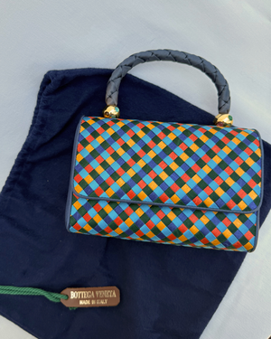 Colorful checkered handbag with braided handle on a navy fabric dust cover on white background