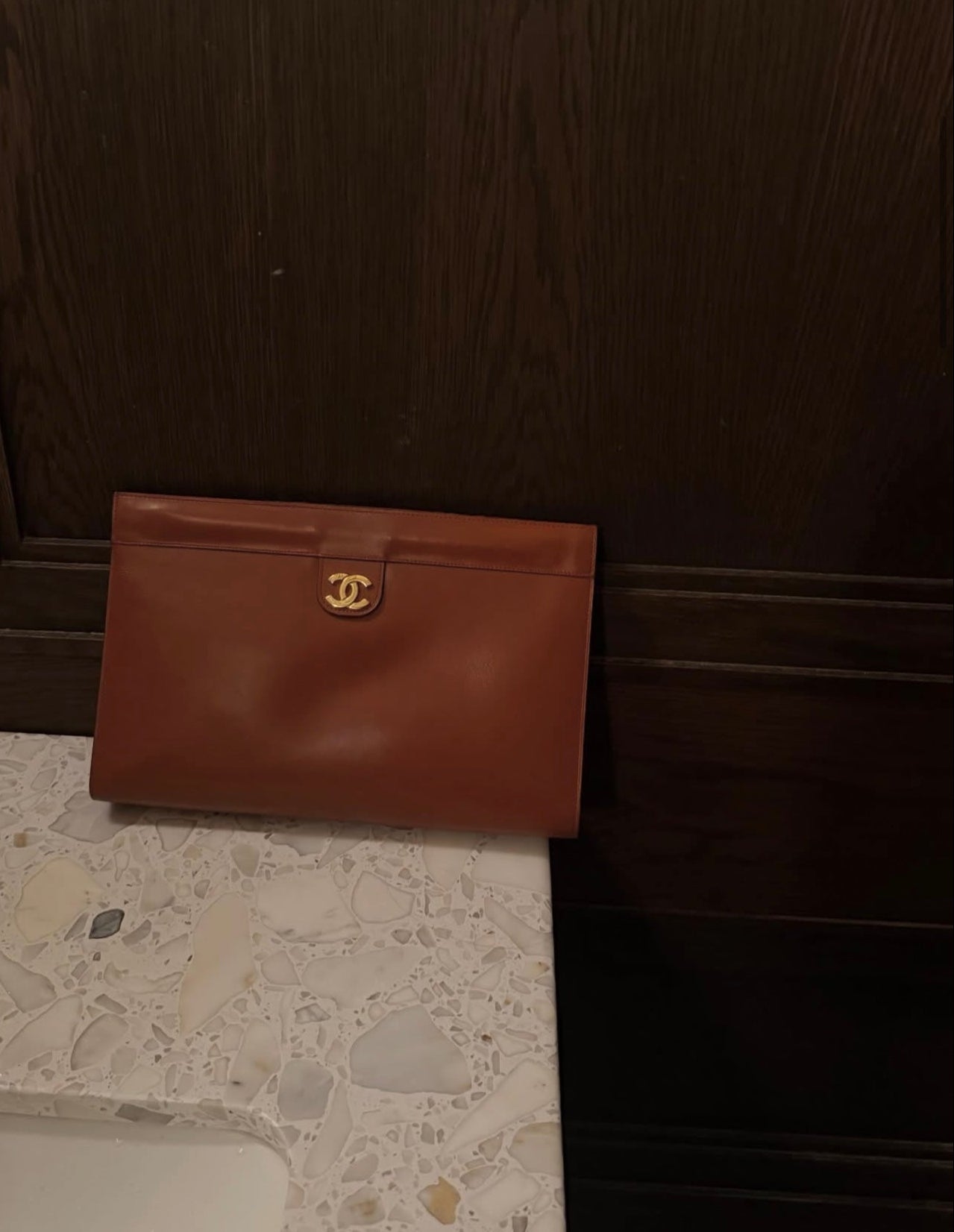 A brown leather clutch bag with a gold-tone interlocking CC logo, resting on a white speckled stone countertop against a dark wood background.