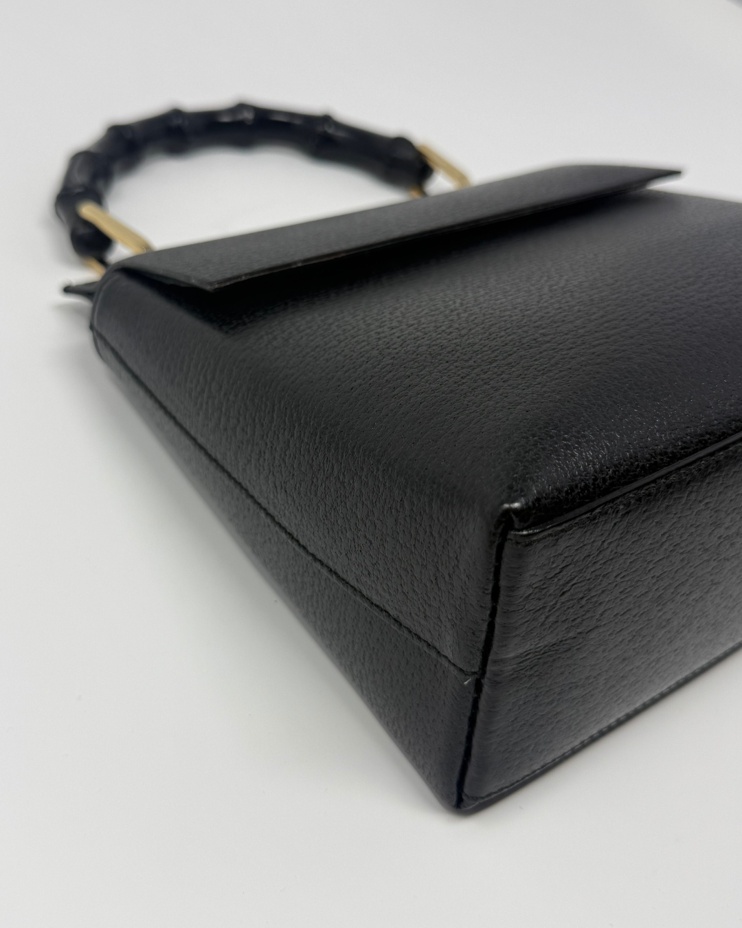 A close-up of a black textured leather handbag with a geometric triangular body and a black woven handle accented by gold-tone hardware, set against a plain white background.