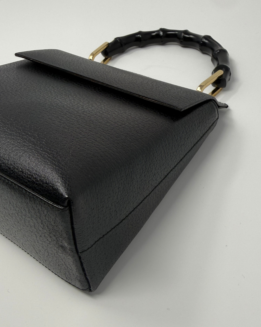 A close-up of a black textured leather handbag with a distinctive curved handle made of black bamboo-like links connected by gold-tone metal hardware, set against a plain light gray background.