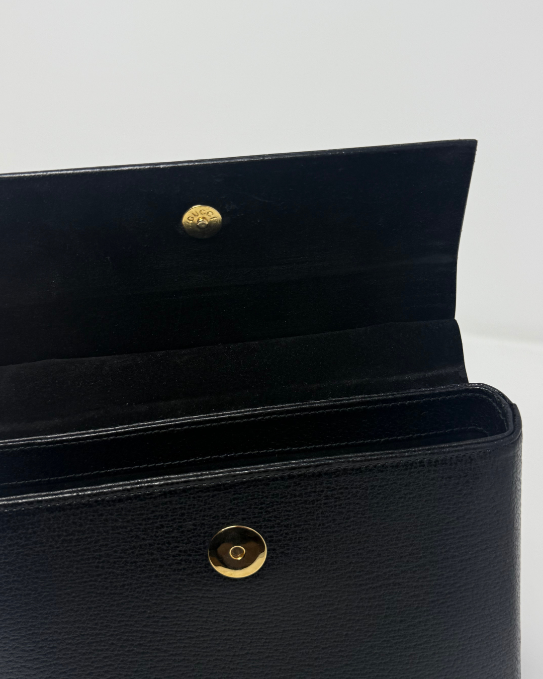 Close-up of a black textured leather clutch or wallet with gold-tone snap buttons, shown open against a plain light background.