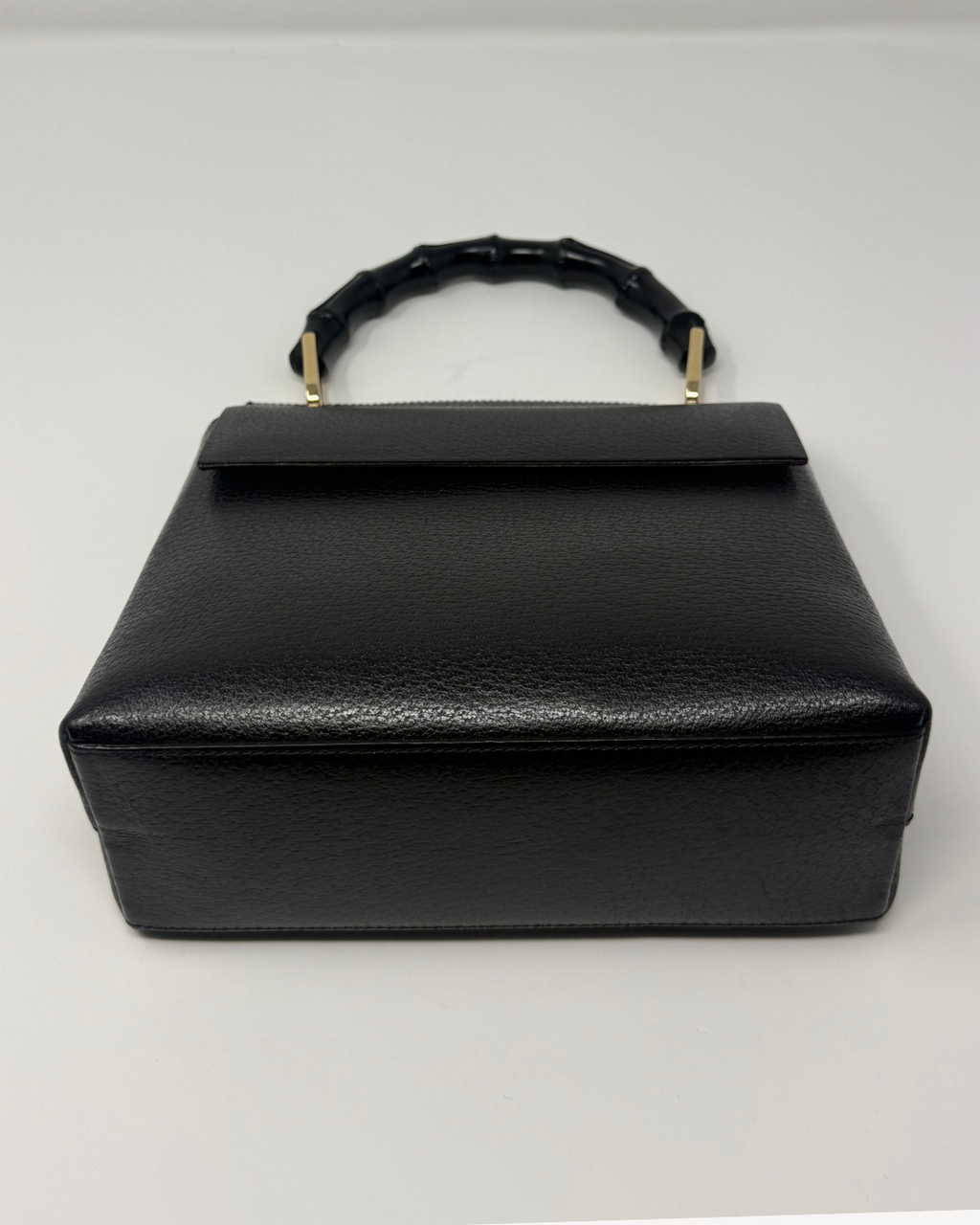 A black textured leather handbag with a structured rectangular shape, a top flap, and a curved handle made of black bamboo-like links with gold-tone metal accents, shown on a plain light background.