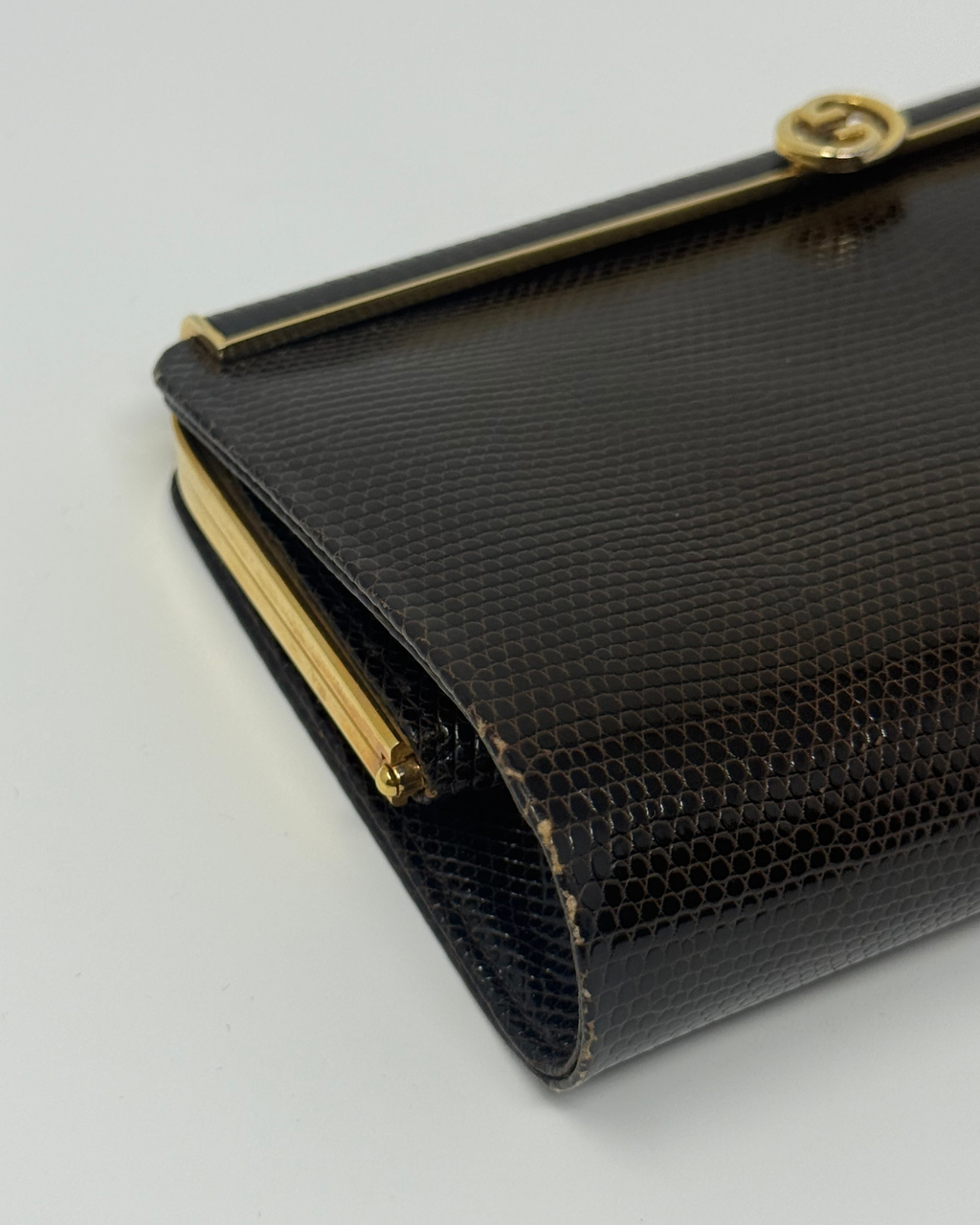 A close-up of a brown lizard python-textured clutch bag with gold-tone hardware and a gold interlocking G logo clasp, indicating the Gucci brand.