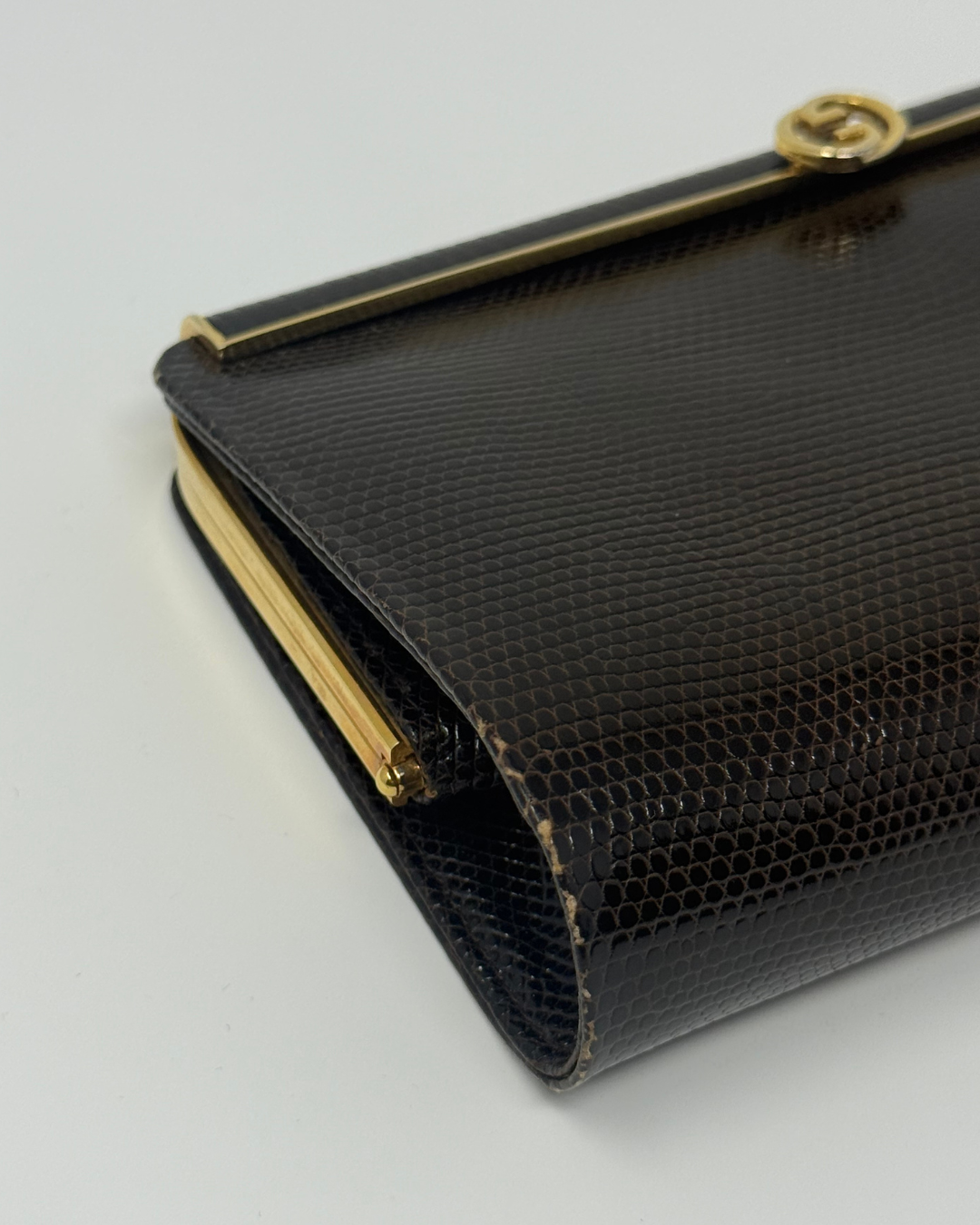 A close-up of a brown lizard python-textured clutch bag with gold-tone hardware and a gold interlocking G logo clasp, indicating the Gucci brand.