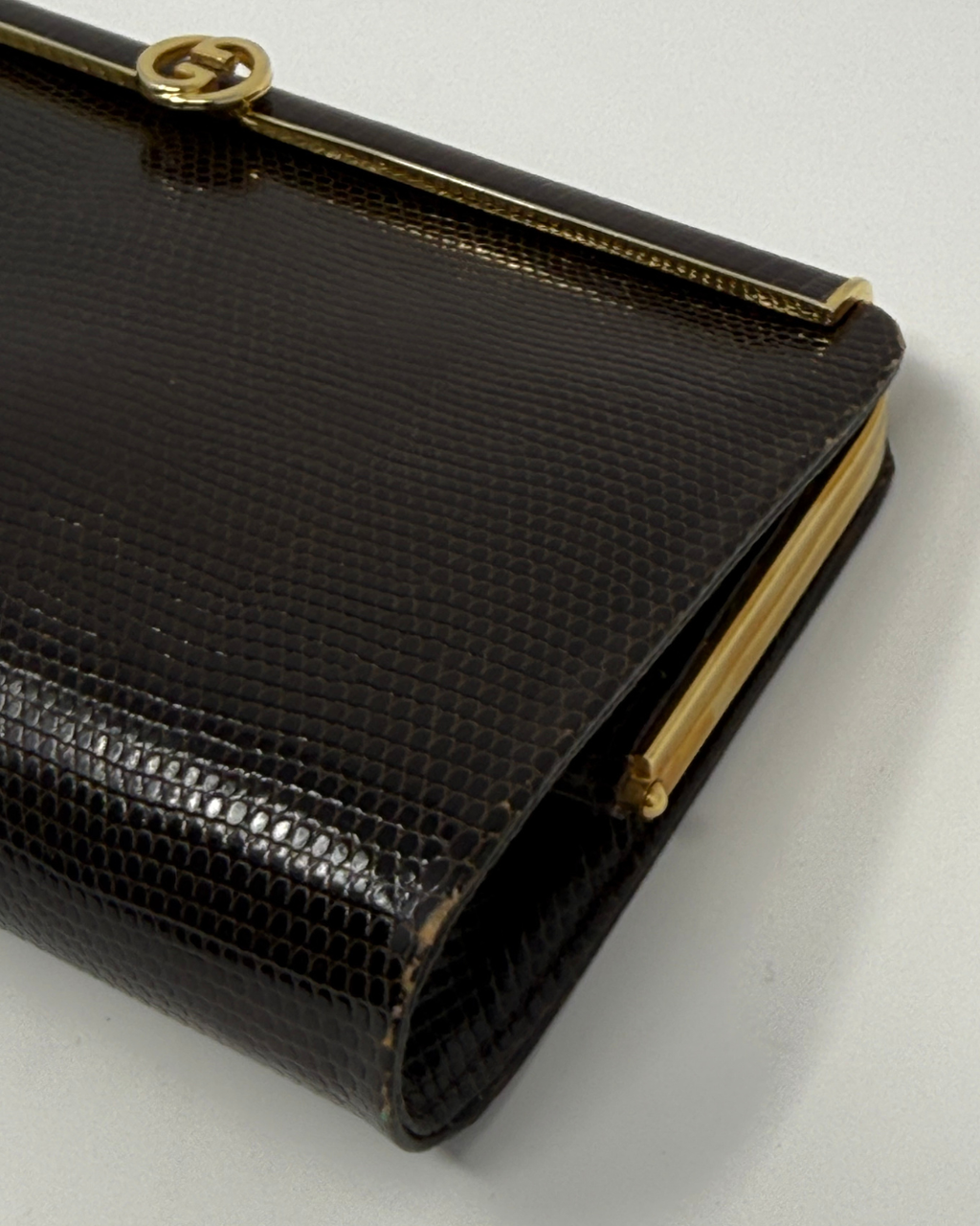 A close-up of a dark brown textured clutch bag with gold-tone hardware and a Gucci interlocking G logo clasp, set against a plain light gray background.