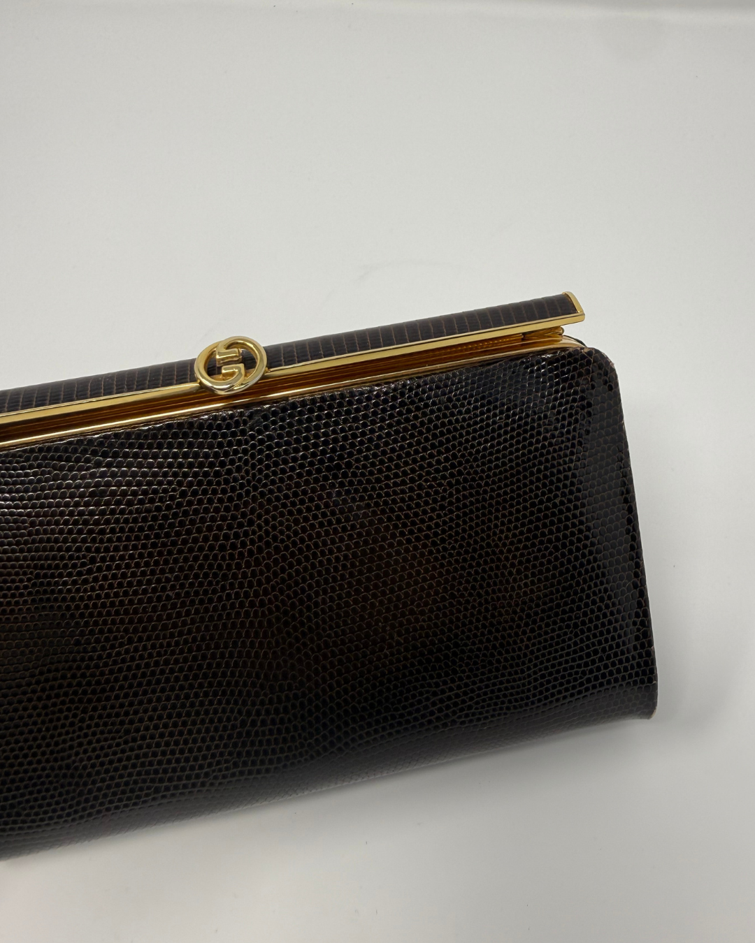 A close-up of a brown textured clutch purse with a gold-tone metal frame and a circular interlocking G logo clasp, indicating the Gucci brand.